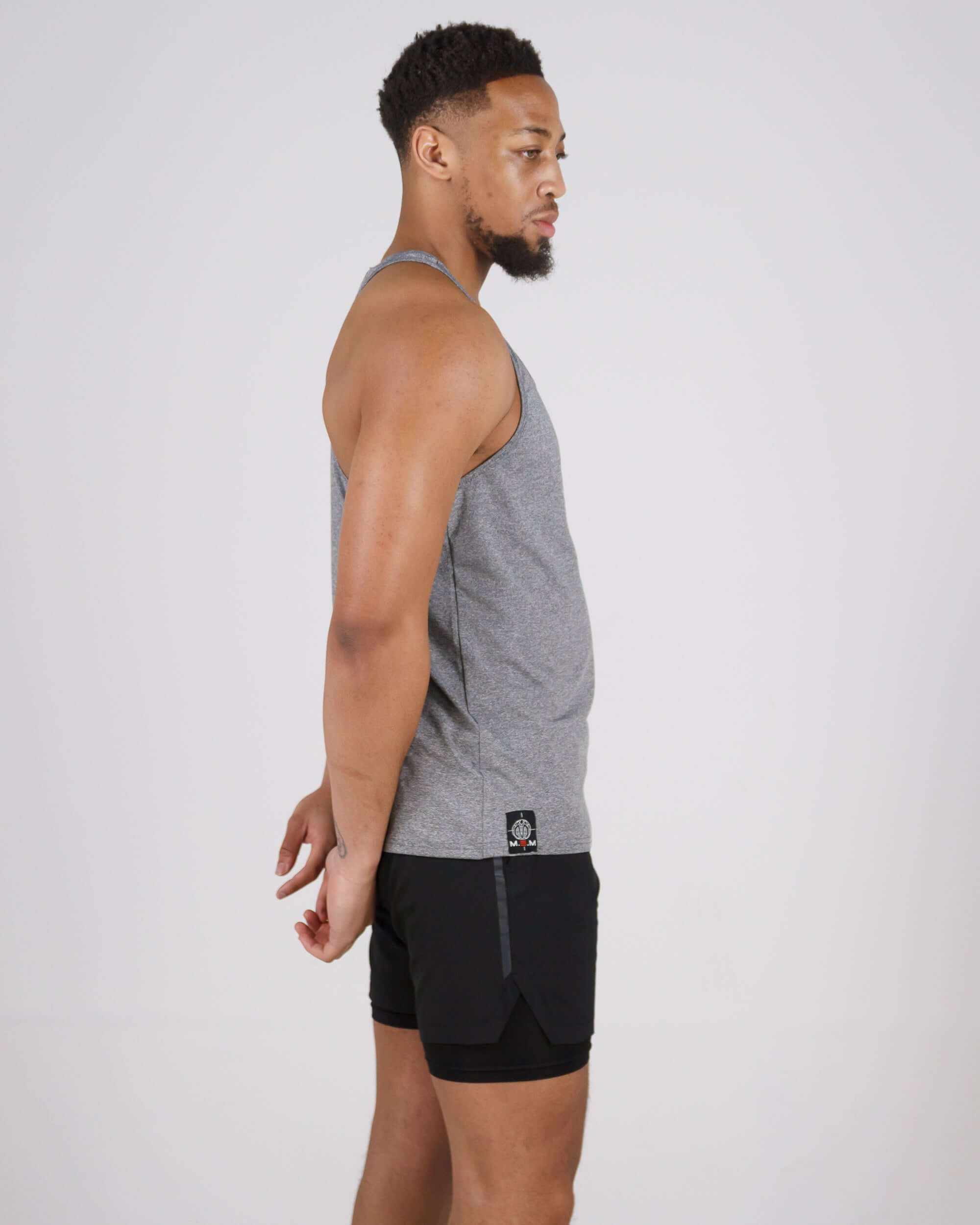 Man wearing grey Pinnacle Slim Fit Tank Top with black shorts, showcasing side profile. Premium polyamide and spandex blend for peak performance.